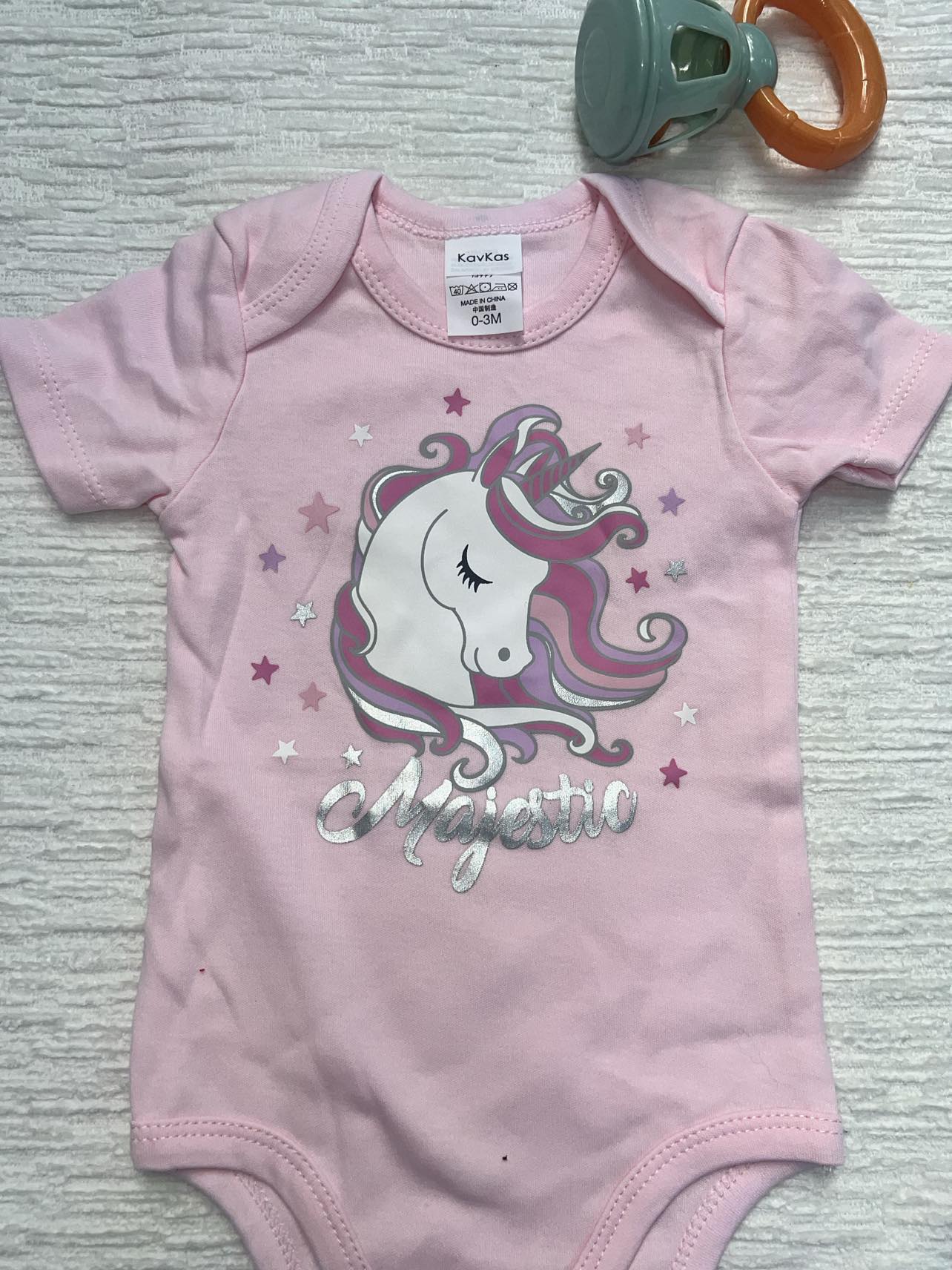 Pink baby onesie with a unicorn with pink, purple, and silver stars and the words: Majestic