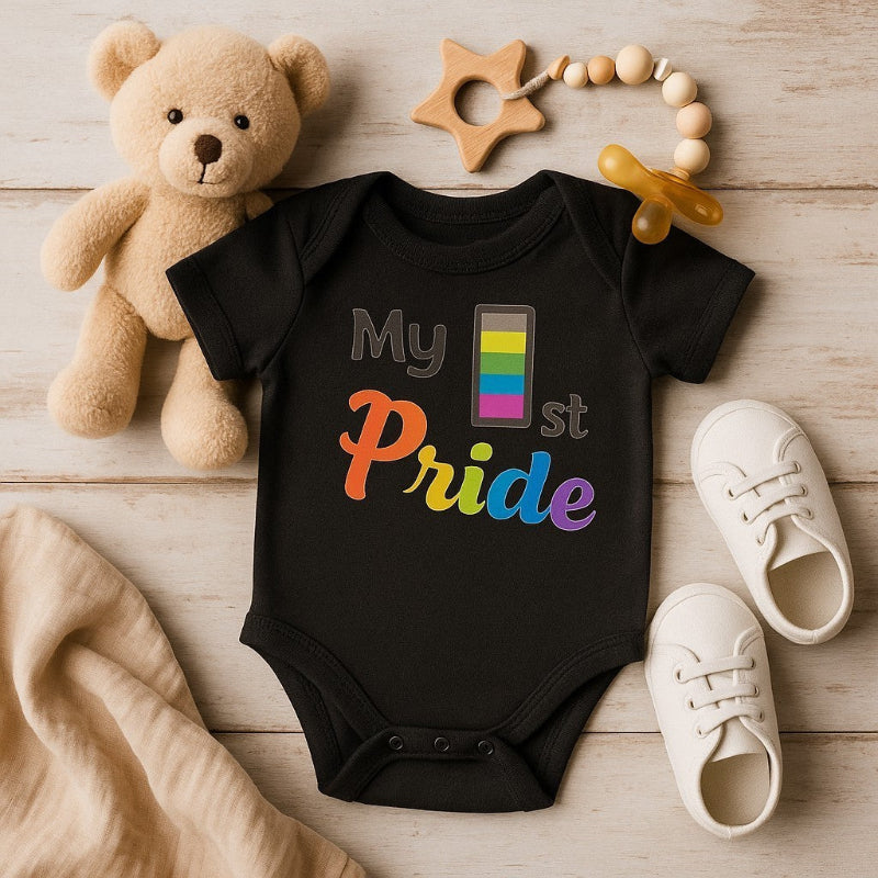 Black baby onesie with rainbow text, teddy bear, shoes, and toys on a wooden surface