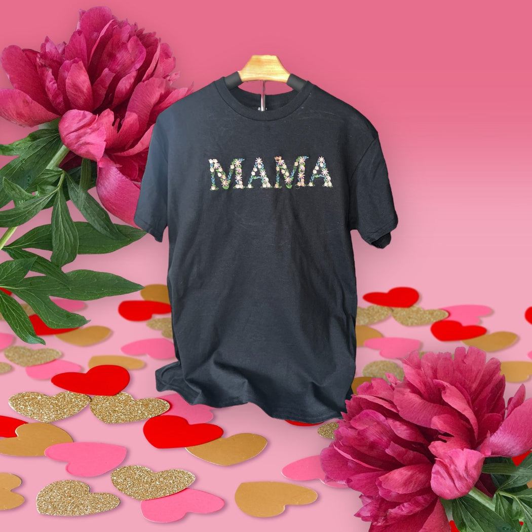 A black t-shirt with the word 'MAMA' written in a floral pattern in the center, displayed on a hanger with floral and heart-shaped decorations around it.