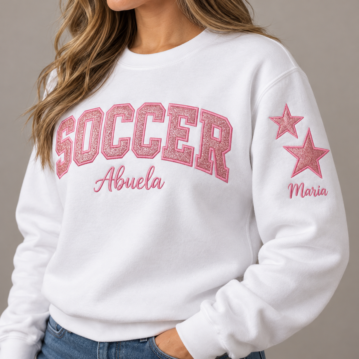 A woman wearing a white crewneck sweatshirt with 'SOCCER Abuela' printed in pink across the chest and two pink stars with the name 'Maria' on the sleeve, paired with blue jeans.