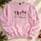 A light pink crewneck sweatshirt with a festive graphic of holiday cocktails and the text 'full of holiday spirit' printed on the chest. The sweatshirt is laid flat on a textured cream fabric background decorated with pine branches and a red ornament.