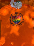 An orange t-shirt with a graphic design supporting LGBTQ+ pride, featuring a heart shape made from different colored arrows forming a rainbow, and the text 'Love is Love'.