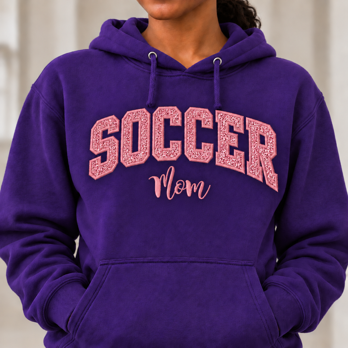 A person wearing a purple pullover hoodie with 'SOCCER Mom' printed on the front in pink. The image is cropped from the chin down, showing the hoodie's drawstrings, front pocket, and textured pink lettering.