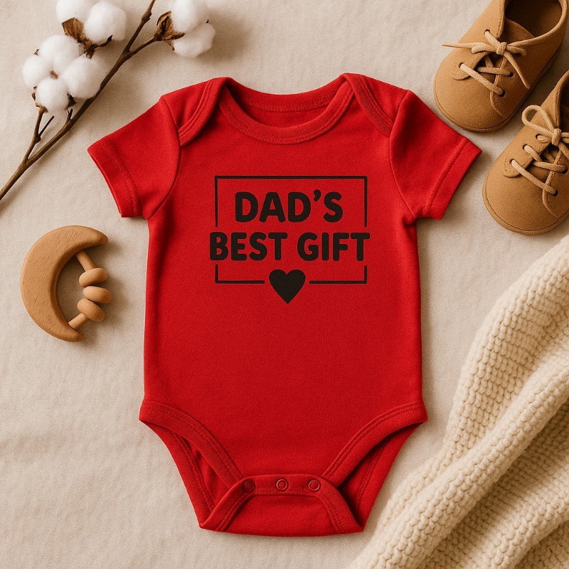Red baby onesie with 'Dad's Best Gift' text on a beige surface with toys and a branch.