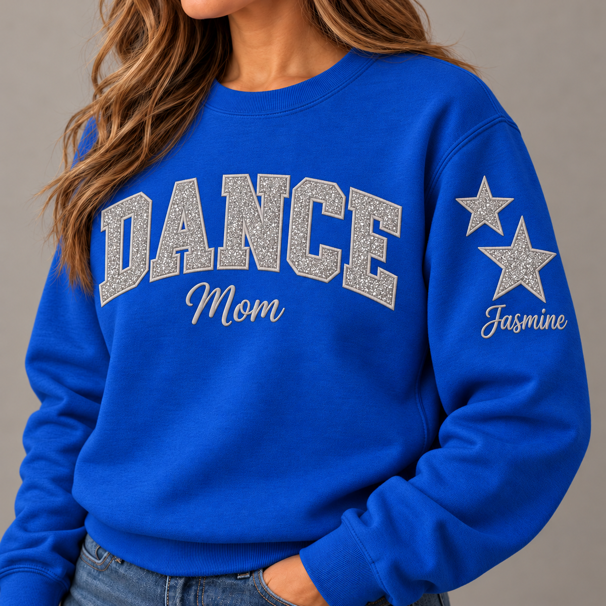 A woman wearing a bright blue crewneck sweatshirt with 'DANCE Mom' printed across the chest in glittery silver letters and 'Jasmine' with two stars on the sleeve, paired with blue jeans against a neutral gray background.