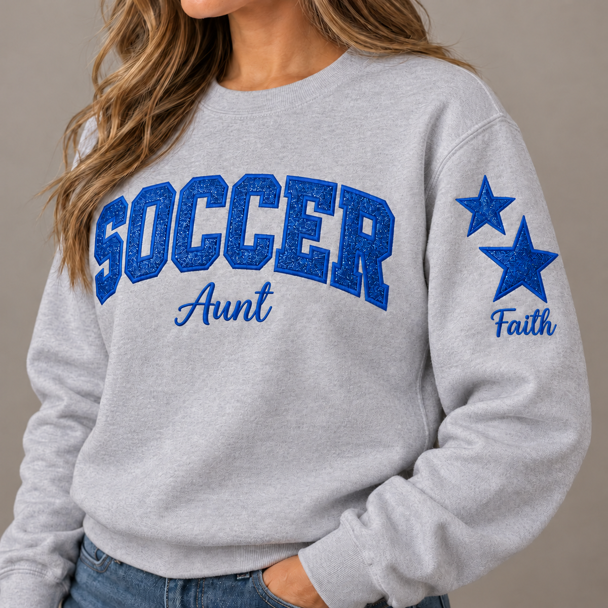 A woman wearing a light heather gray crewneck sweatshirt with 'SOCCER Aunt' printed in blue across the chest and 'Faith' with two blue stars on the sleeve, paired with blue jeans.