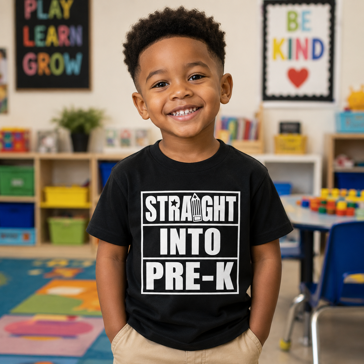 A young boy smiling in a colorful children's classroom, wearing a black t-shirt with a bold white graphic that reads 'STRAIGHT INTO PRE-K'.