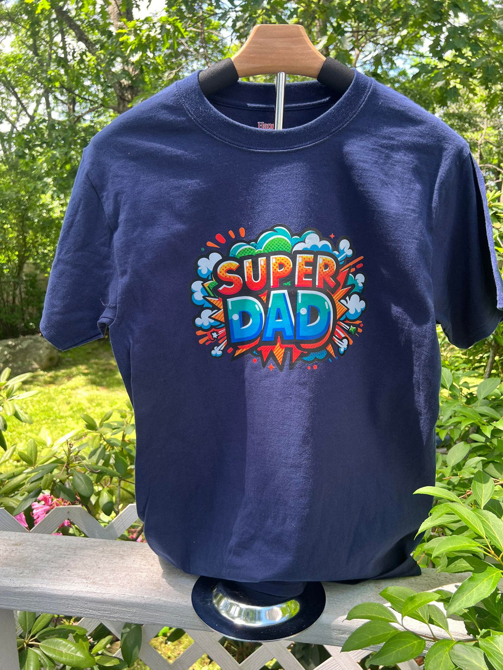 A navy blue t-shirt with a graphic print that reads 'Super Dad' in colorful letters, displayed on a wooden hanger outdoors with foliage in the background.