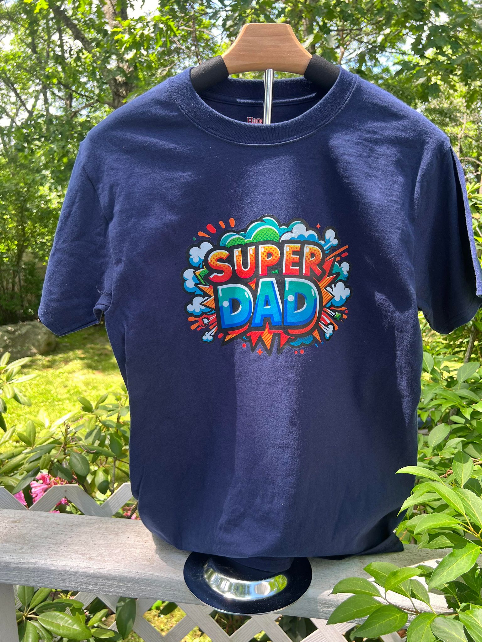 A navy blue t-shirt with a graphic print that reads 'Super Dad' in colorful letters, displayed on a wooden hanger outdoors with foliage in the background.