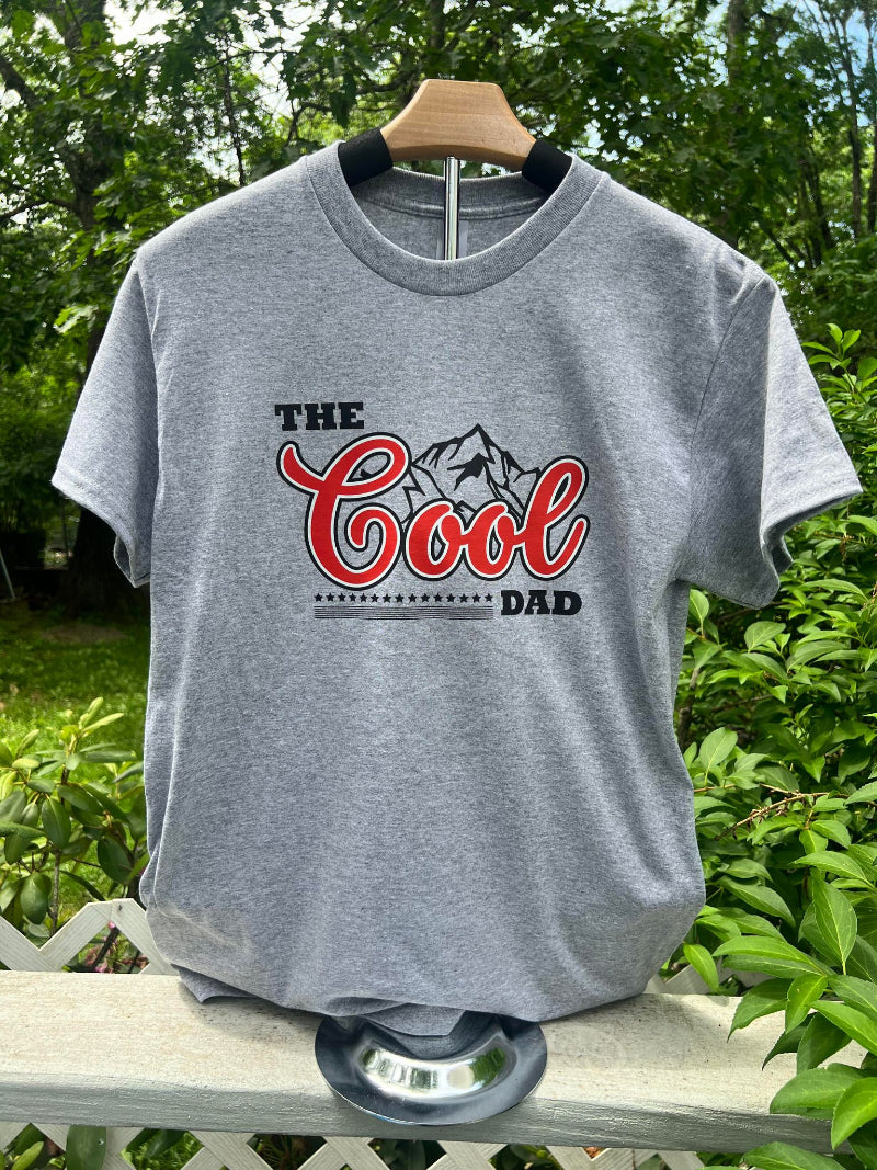 A gray t-shirt with 'The Cool Dad' text and a graphic of a beer bottle on it, displayed on a wooden hanger outdoors.