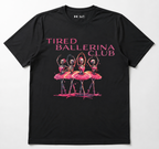 A black short-sleeve t-shirt with a graphic print of four ballerinas in pink tutus under the text 'TIRED BALLERINA CLUB' in pink, on a light gray background.
