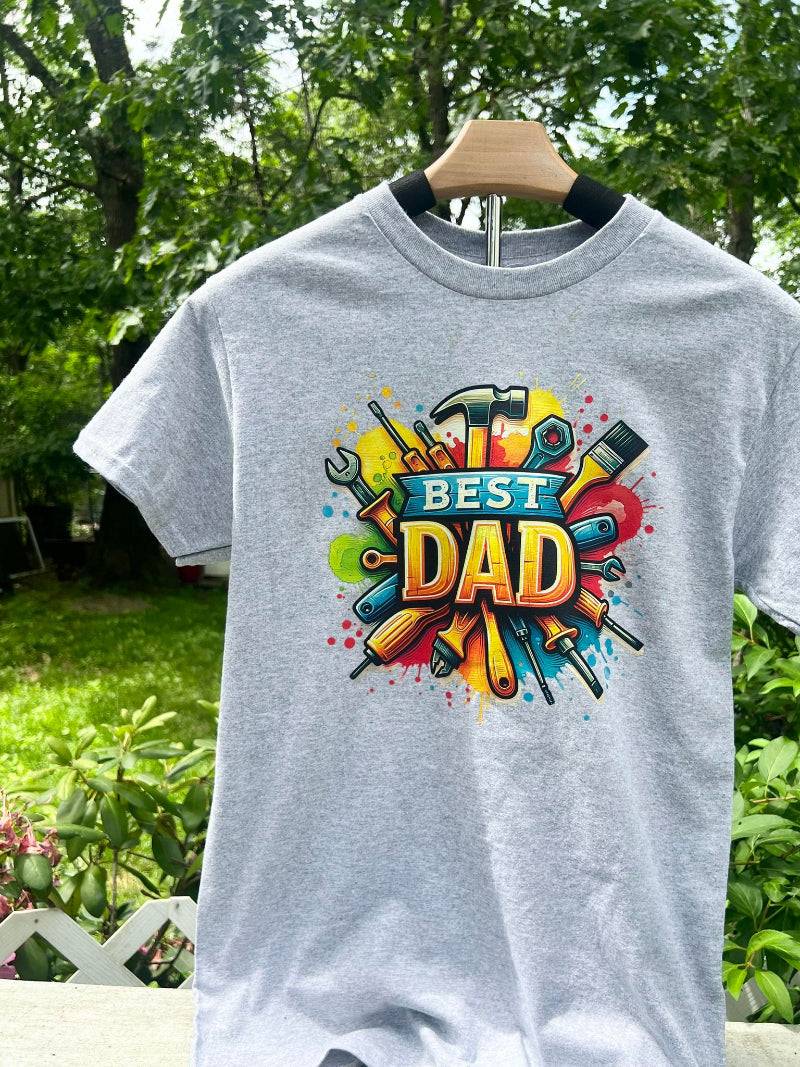 A gray t-shirt with a colorful graphic print that reads 'BEST DAD' surrounded by various tools, displayed on a wooden hanger outdoors.