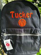A black personalized backpack with orange embroidered text reading 'Tucker' and an orange basketball graphic, shown outdoors on a wooden fence with green foliage in the background. A white 'Design Wealthy' hangtag is attached near the front pocket.