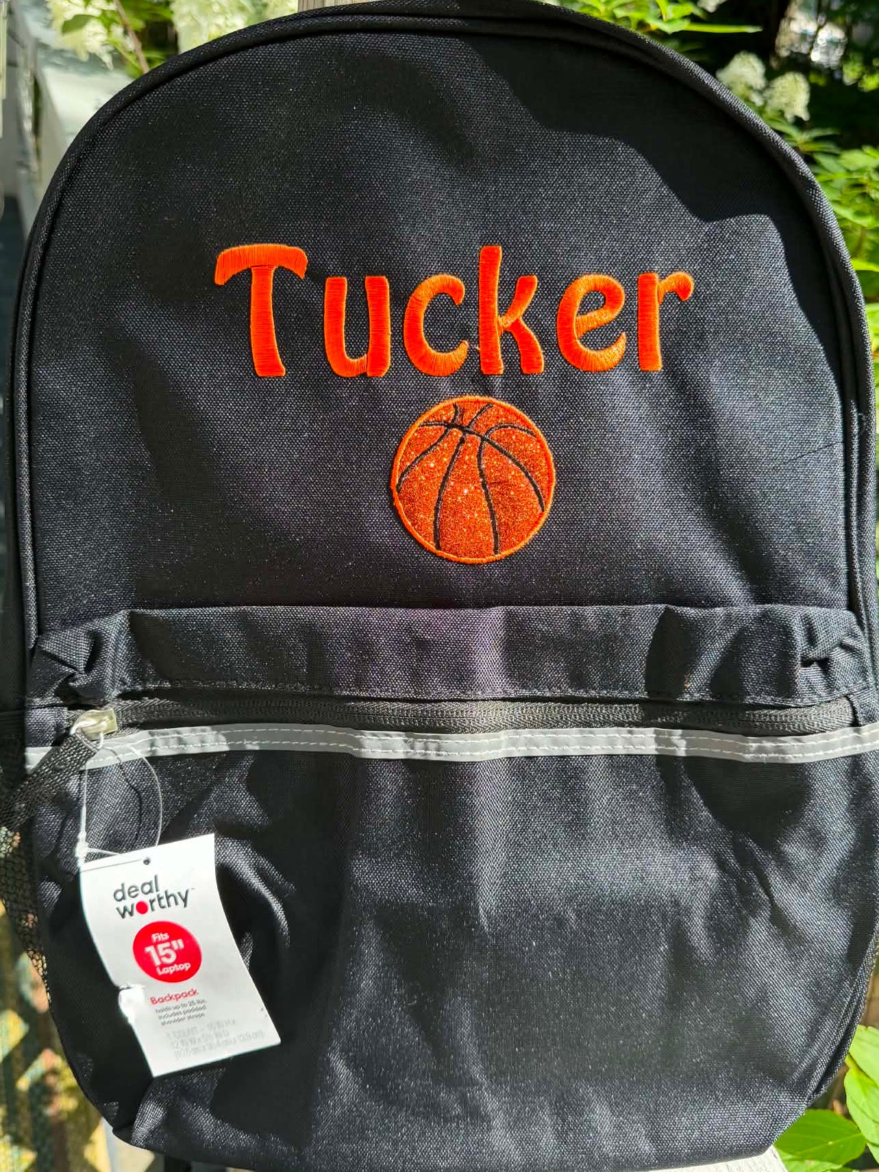 A black personalized backpack with orange embroidered text reading 'Tucker' and an orange basketball graphic, shown outdoors on a wooden fence with green foliage in the background. A white 'Design Wealthy' hangtag is attached near the front pocket.