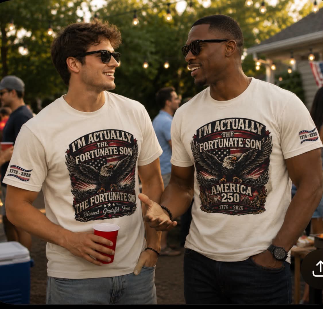 Two men wearing matching white graphic t-shirts with a distressed black and red eagle design that reads 'I'M ACTUALLY THE FORTUNATE SON' and 'AMERICA 250'. The photo is taken at an outdoor evening gathering with people and string lights in the background.