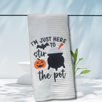 White Halloween towel embroidered with a cauldron, pumpkin, bat, and the words “I’m Just Here to Stir the Pot.”
