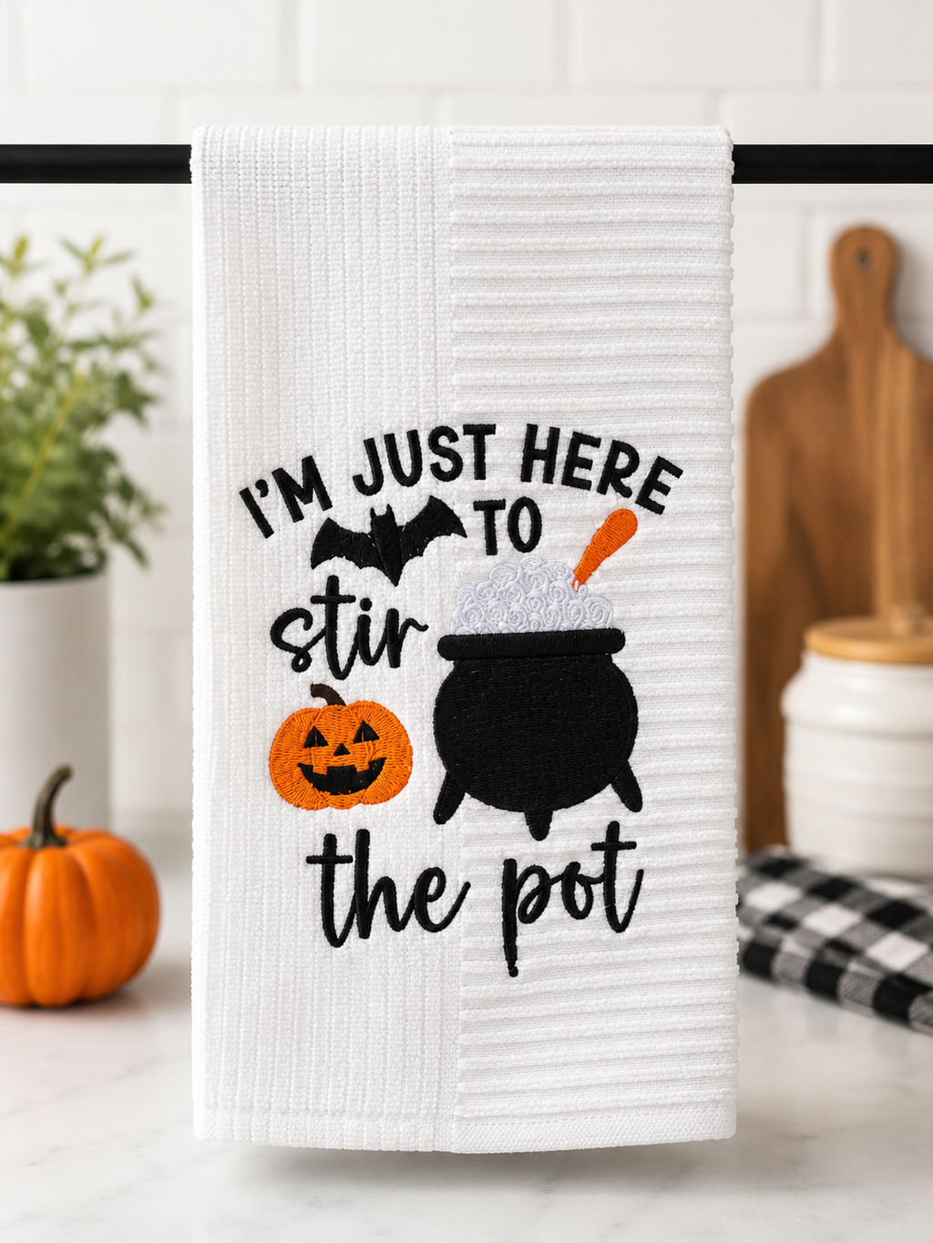 White embroidered Halloween kitchen towel featuring the phrase "I'm Just Here to Stir the Pot" with a black cauldron, smiling jack-o'-lantern, bat, and orange accents, displayed in a modern kitchen