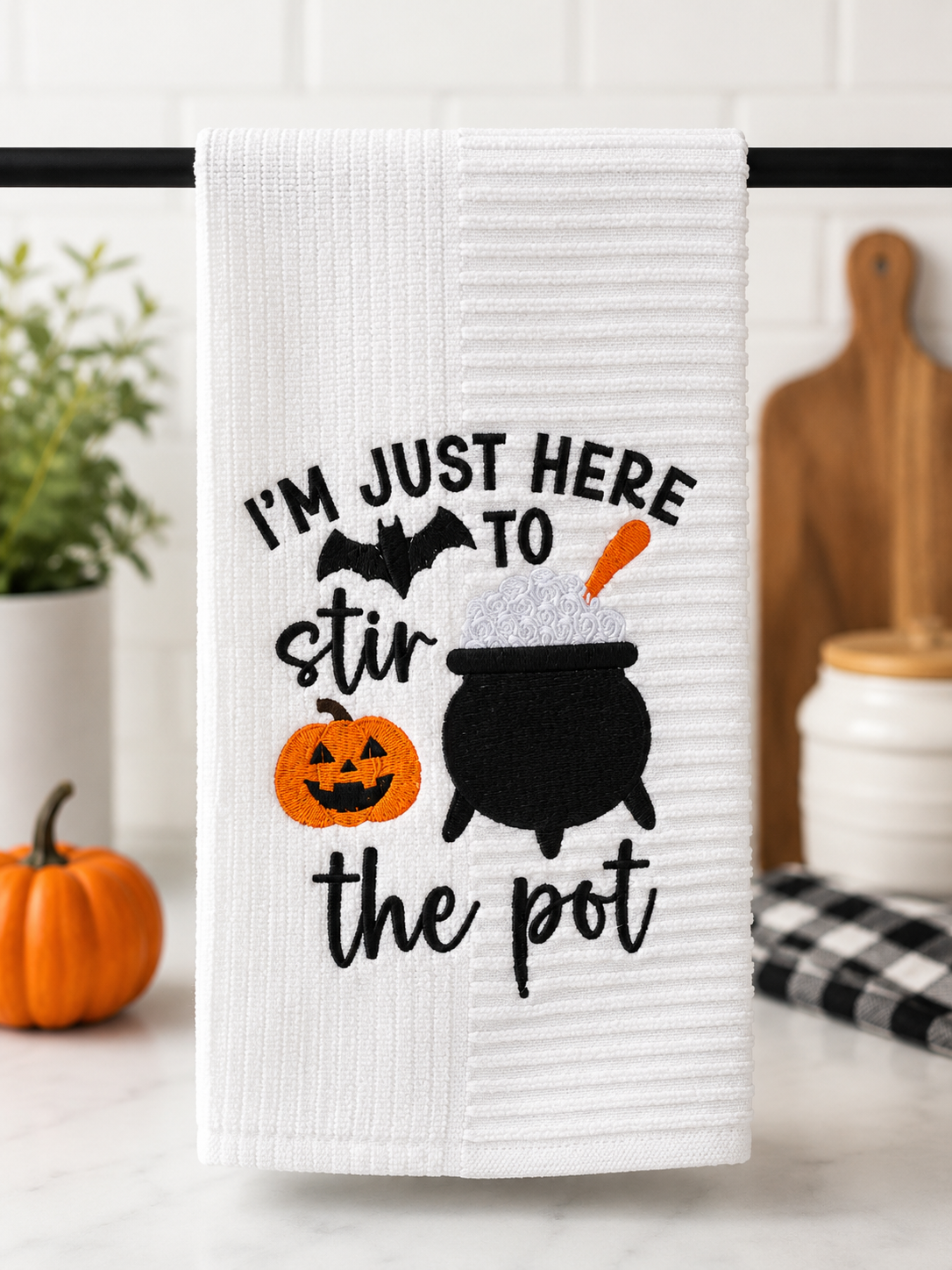 White embroidered Halloween kitchen towel featuring the phrase "I'm Just Here to Stir the Pot" with a black cauldron, smiling jack-o'-lantern, bat, and orange accents, displayed in a modern kitchen