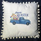 A beige embroidered throw pillow with a vintage blue truck filled with spring flowers and the text 'Happy Easter' embroidered on it, surrounded by a pom-pom trim.