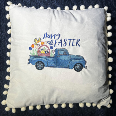 A beige embroidered throw pillow with a vintage blue truck filled with spring flowers and the text 'Happy Easter' embroidered on it, surrounded by a pom-pom trim.