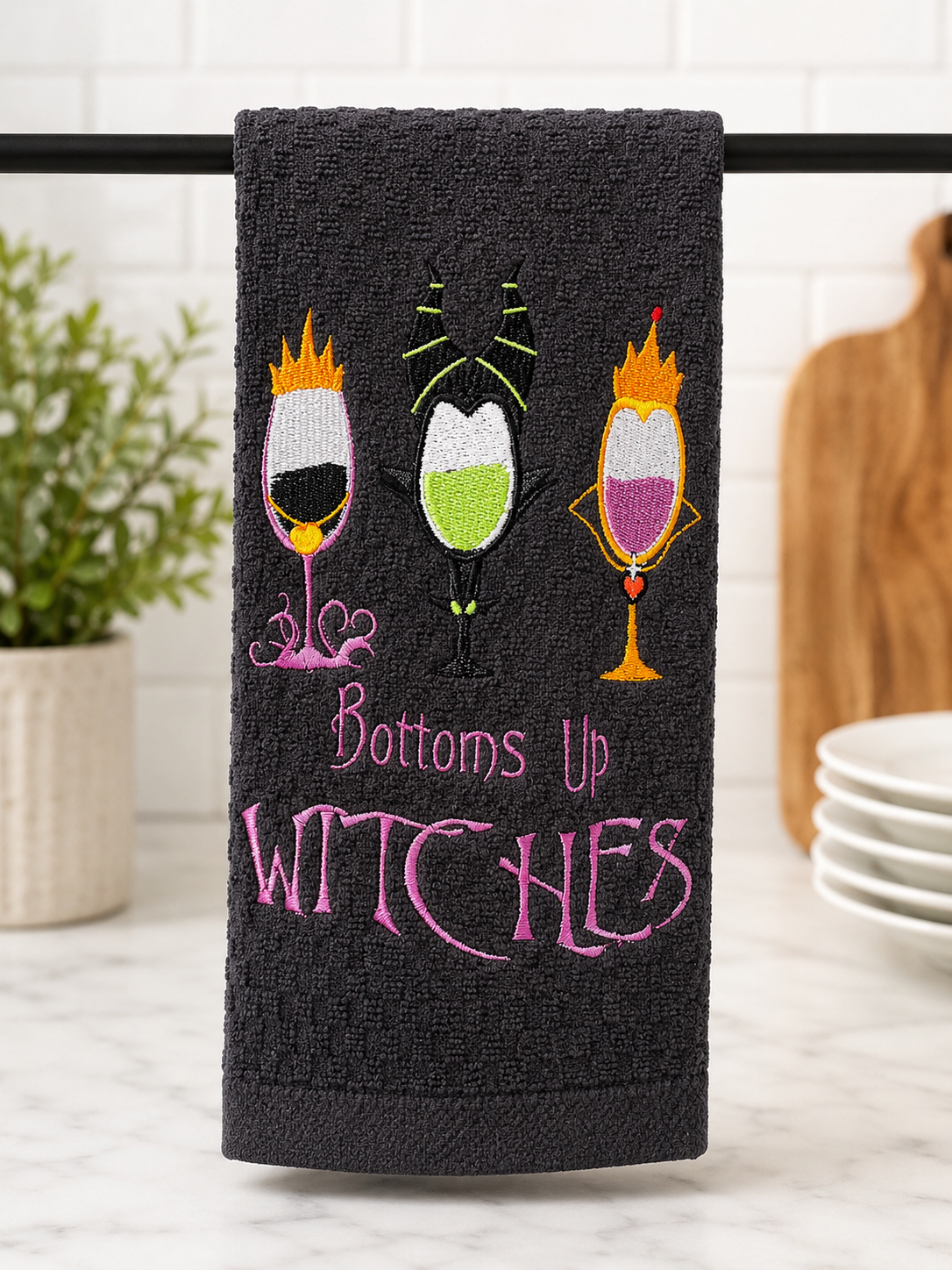 Black embroidered Halloween kitchen towel featuring the phrase "Bottoms Up Witches" with three spooky wine glasses inspired by witches, displayed in a modern farmhouse kitchen.