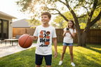 A boy wearing a white graphic t-shirt with a basketball design stands in a sunny backyard holding a basketball, while a girl in the background takes a photo with her phone.