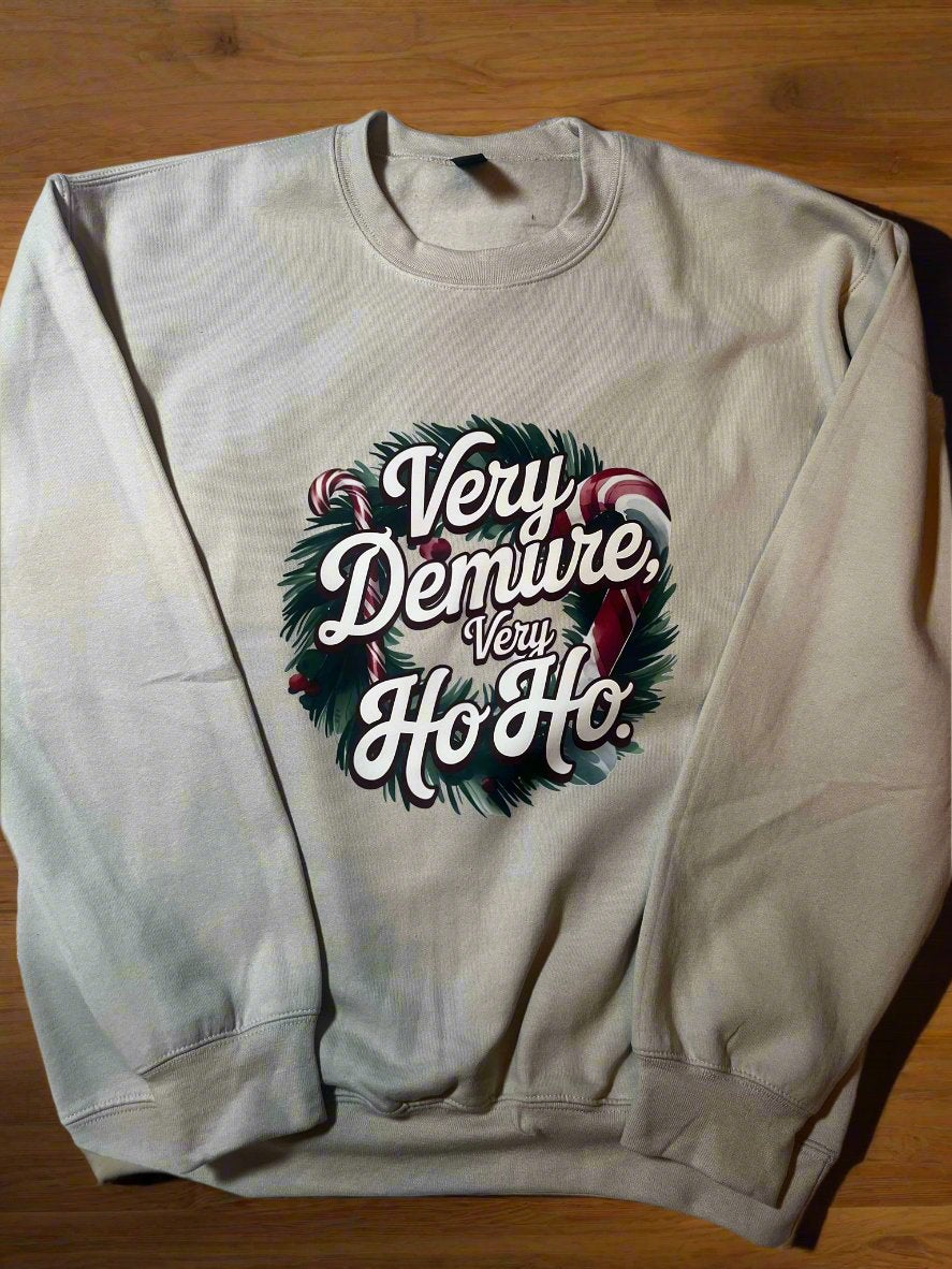 A cream-colored sweatshirt with 'Very Demure, Very Ho-Ho' text and a Christmas wreath design, displayed on a hanger.