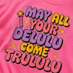 A close-up of a bright pink t-shirt featuring a colorful graphic print with the text 'MAY ALL YOUR DELULU COME TRULULU' in purple, pink, and yellow bubble letters with black outlines, accompanied by small yellow stars and a wand illustration.