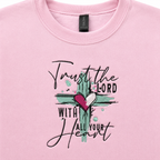 A close-up of a light pink crewneck sweatshirt featuring a embroidered design with the text 'Trust the LORD WITH ALL YOUR Heart' and a cross design with a pink heart.