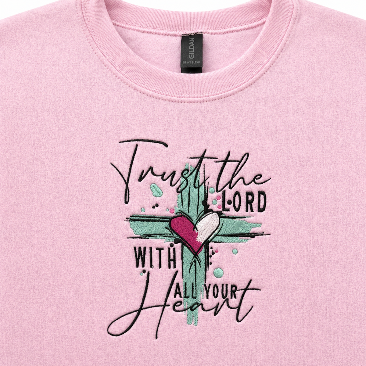 A close-up of a light pink crewneck sweatshirt featuring a embroidered design with the text 'Trust the LORD WITH ALL YOUR Heart' and a cross design with a pink heart.