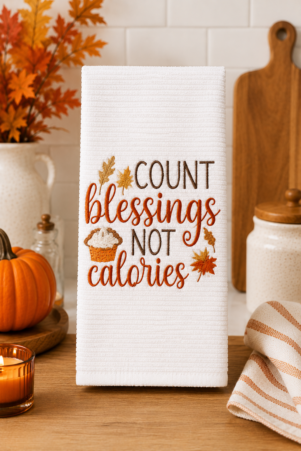 White embroidered kitchen towel featuring the phrase "Count Blessings Not Calories" with autumn leaves and pumpkin pie, displayed in a cozy farmhouse kitchen decorated for fall.
