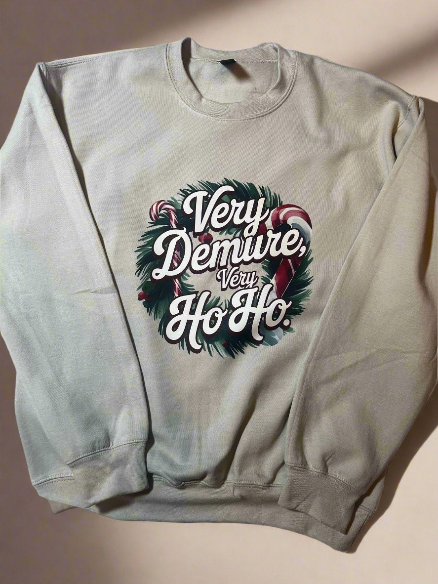 A cream-colored sweatshirt with 'Very Demure, Very Ho-Ho' text and a Christmas wreath design, laid out