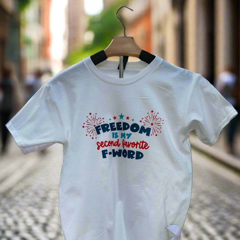 White graphic t-shirt with red, white, and blue fireworks design and the funny quote “Freedom Is My Second Favorite F-Word” hanging outside on a hanger. 