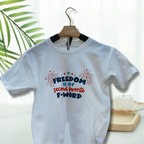 White graphic t-shirt with red, white, and blue fireworks design and the funny quote “Freedom Is My Second Favorite F-Word” hanging outside on a hanger. 