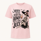 A light pink short-sleeve t-shirt with a graphic print of a skeleton witch holding an orange drink, next to the text 'WHEN YOU'RE READY FOR HALLOWEEN BUT IT'S ONLY JULY'.
