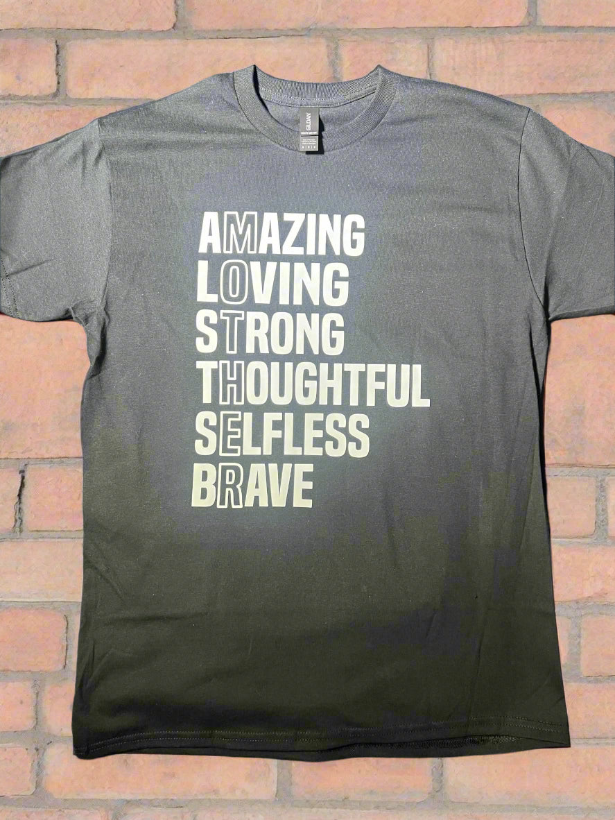 A black short-sleeve graphic tee with white text that reads 'AMAZING LOVING STRONG THOUGHTFUL SELFLESS BRAVE' vertically, with the word 'MOTHER' highlighted at the end with a brick background