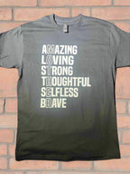 A black short-sleeve graphic tee with white text that reads 'AMAZING LOVING STRONG THOUGHTFUL SELFLESS BRAVE' vertically, with the word 'MOTHER' highlighted at the end with a brick background