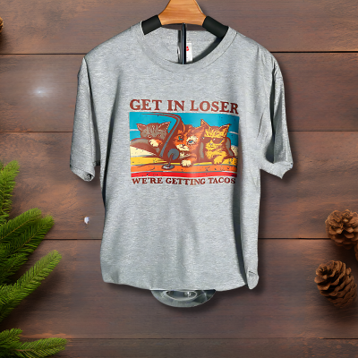 A gray t-shirt with a graphic print of three stylized cats and the text 'GET IN LOSER - WE'RE GETTING TACOS' on it, displayed on a hanger with a variety of food items in the background.