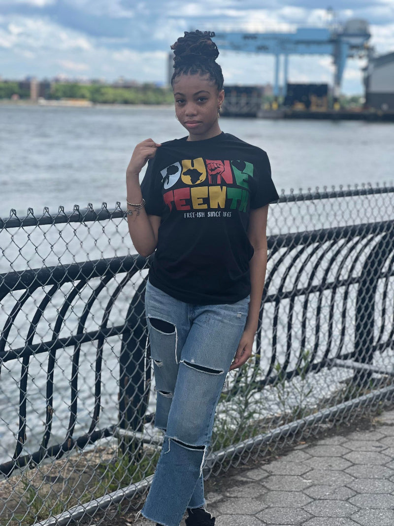 Person wearing a black t-shirt with colorful text, standing by a waterfront.