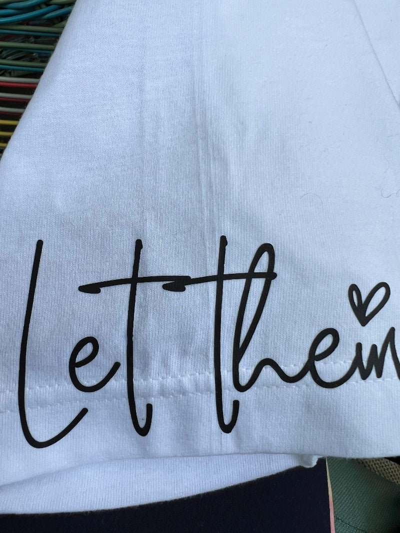Close-up of sleeve design with “Let Them” text and heart graphic on white cotton t-shirt.
