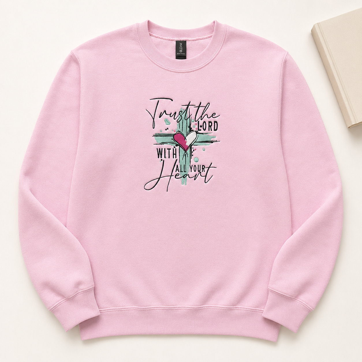 A light pink crewneck sweatshirt featuring a embroidered design  on the chest with a cross, a pink heart, and the text 'Trust the LORD with Your Heart'. The sweatshirt is laid flat on a light beige background with a book corner visible in the top right.