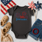 Black baby onesie with 'little firecracker' design, surrounded by American flag, shoes, denim shorts, and decorative items on a light wooden surface.