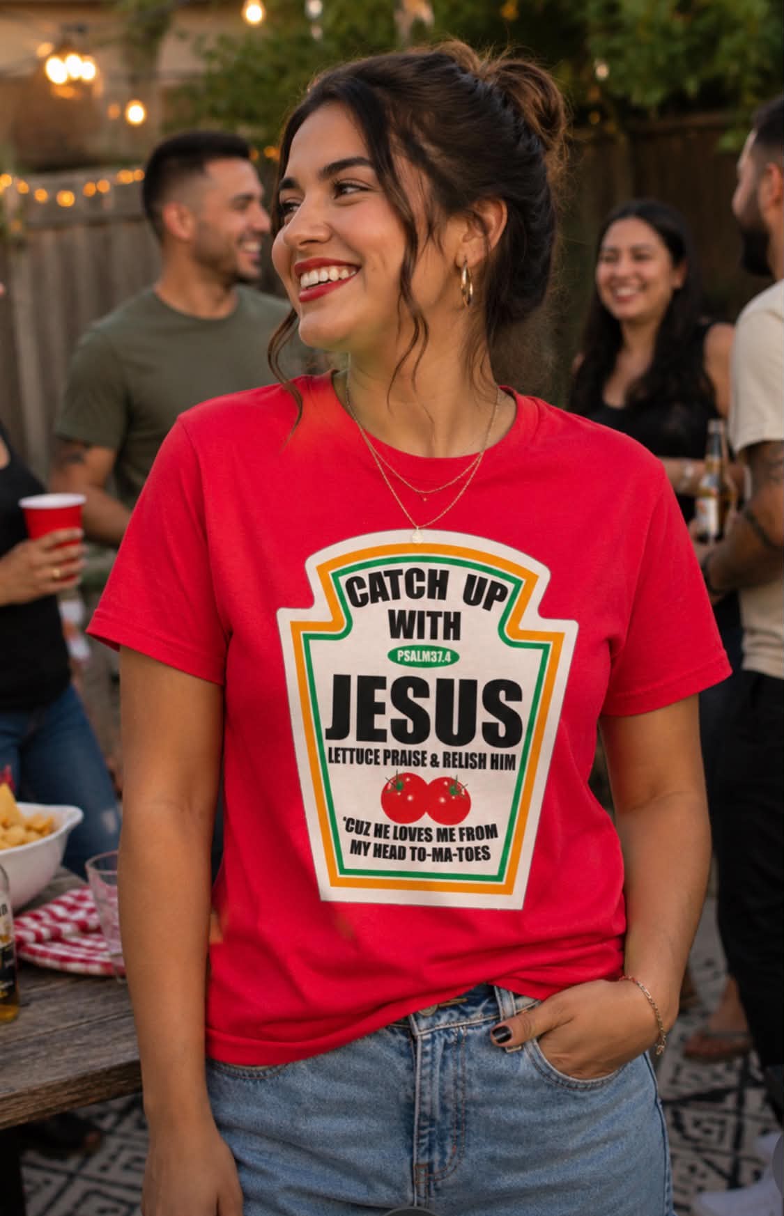 A woman wearing a bright red graphic t-shirt with a ketchup-style label design that reads 'Catch Up With JESUS' and 'LETTE PRAISE ME FROM MY HEAD TO MY TOES' with two tomatoes, photographed at an outdoor party.