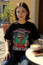 Woman wearing a black t-shirt with a colorful graphic design, sitting outdoors with a cup of coffee.
