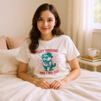 Woman wearing a t-shirt with a graphic design and text, sitting on a bed.