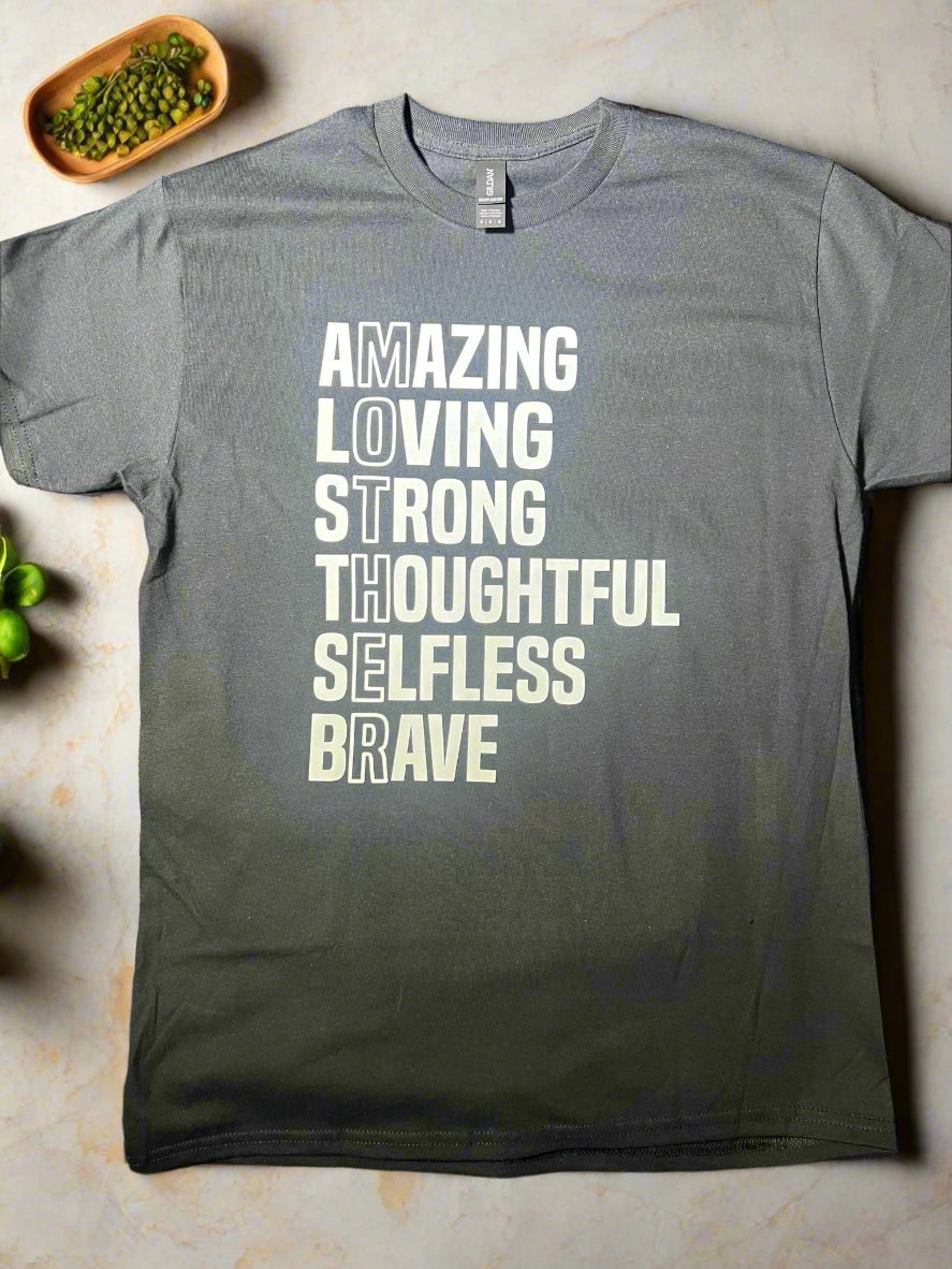 A black short-sleeve graphic tee with white text that reads 'AMAZING LOVING STRONG THOUGHTFUL SELFLESS BRAVE' vertically, with the word 'MOTHER' highlighted at the end.