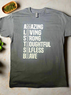 A black short-sleeve graphic tee with white text that reads 'AMAZING LOVING STRONG THOUGHTFUL SELFLESS BRAVE' vertically, with the word 'MOTHER' highlighted at the end.