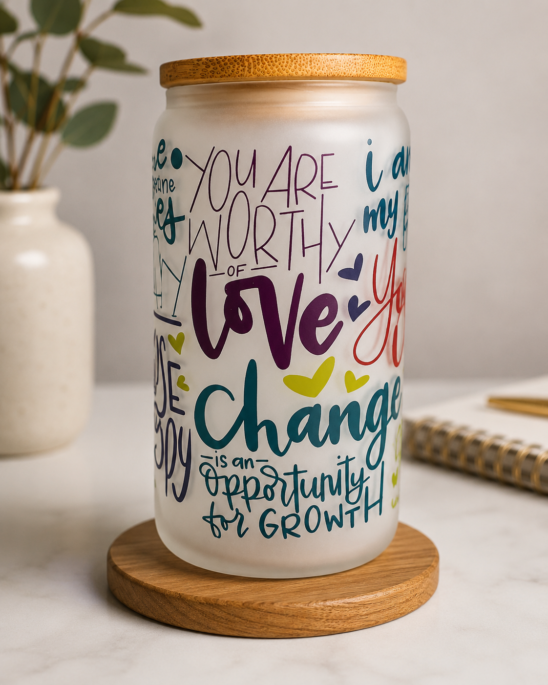 Frosted glass can with a bamboo lid featuring colorful inspirational affirmations including "You Are Worthy of Love" and "Change Is an Opportunity for Growth," displayed in a bright modern home setting.