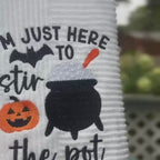 Video of a white Halloween towel embroidered with a cauldron, pumpkin, bat, and the words “I’m Just Here to Stir the Pot.”