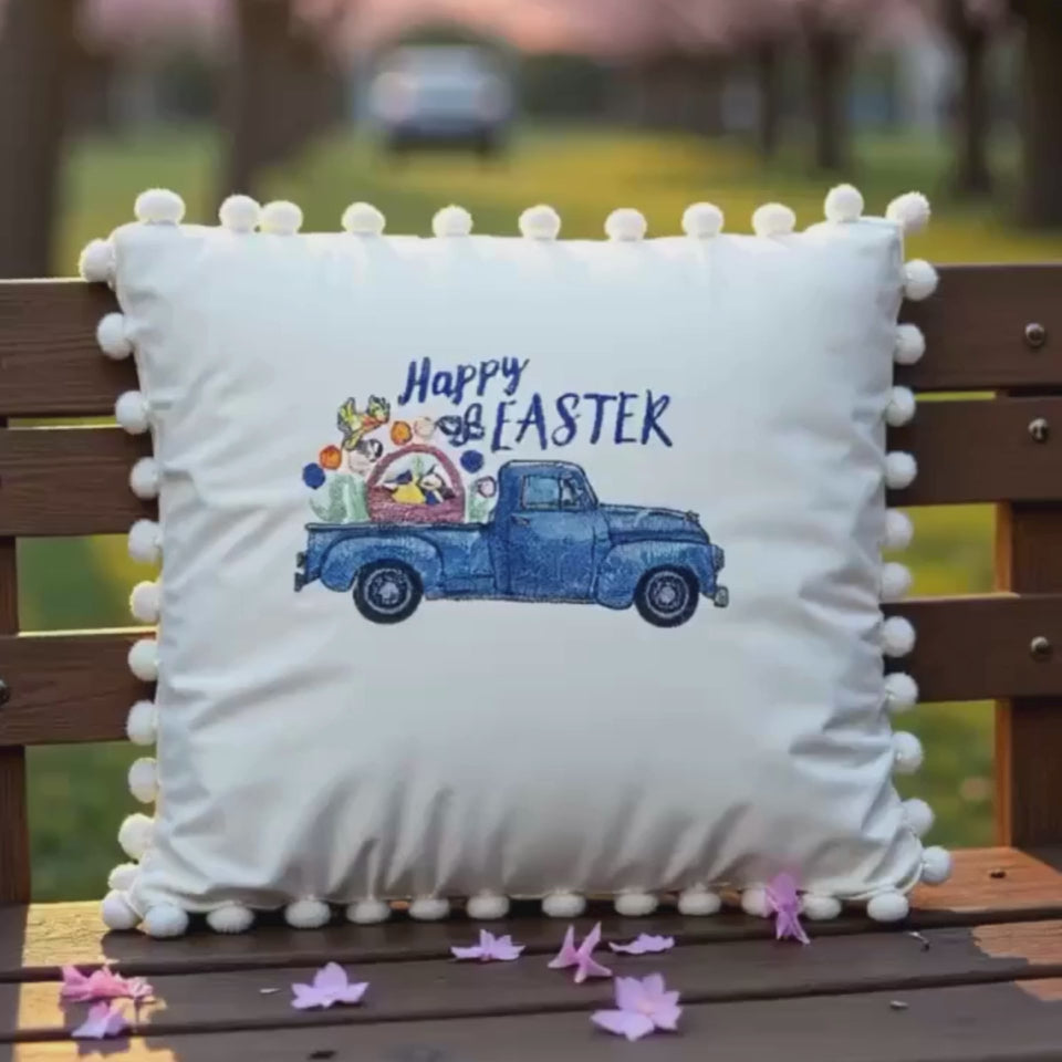 video of beige embroidered throw pillow with a vintage blue truck filled with spring flowers and the text 'Happy Easter' embroidered on it, surrounded by a pom-pom trim.
